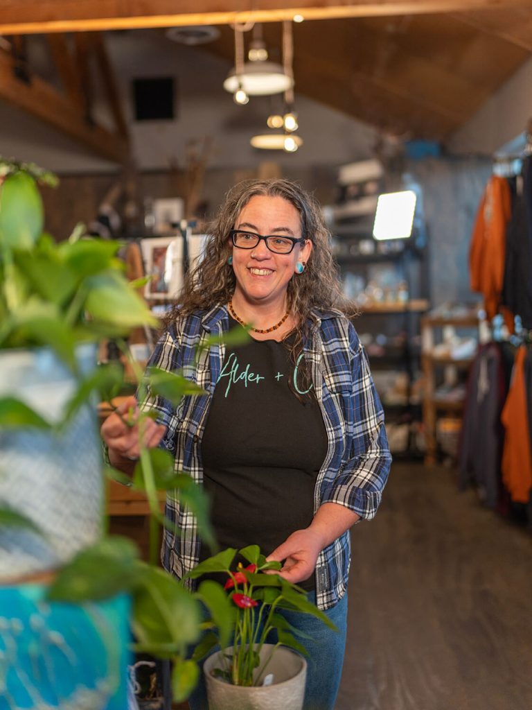 An employee looking at plants inside of Alder + Co. and racks of clothing and goods displayed on shelves in the background.