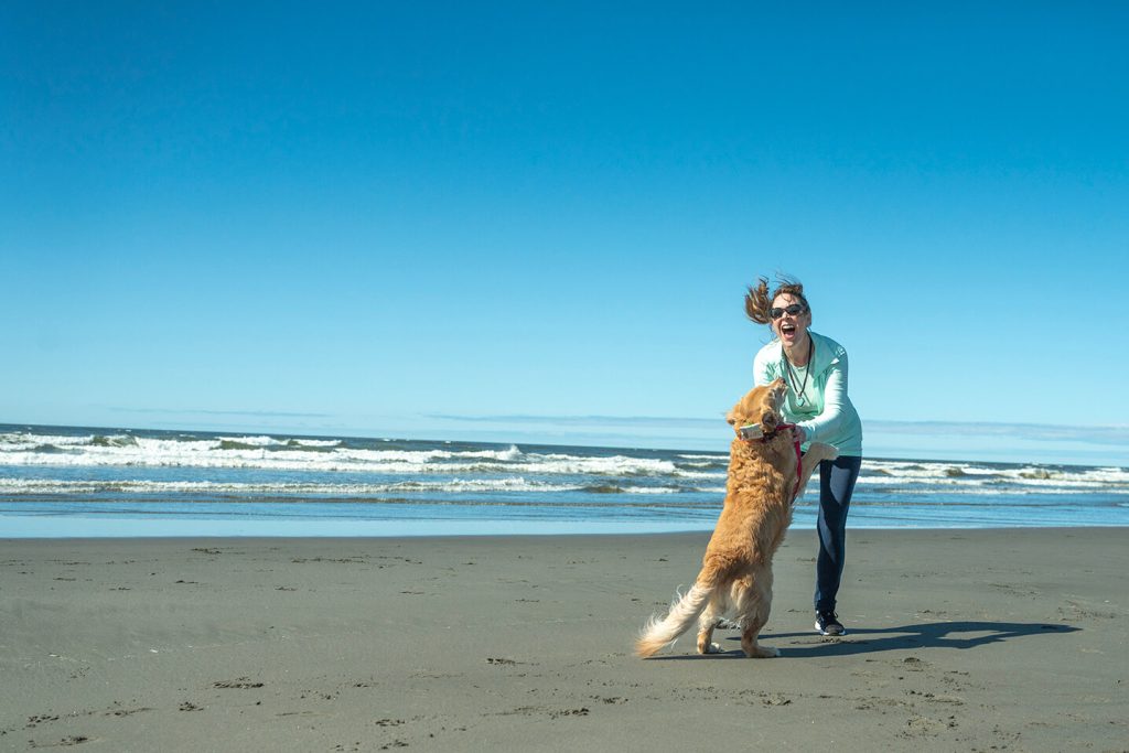 A woman playing with her dog on the beach and crashing waves in the background.