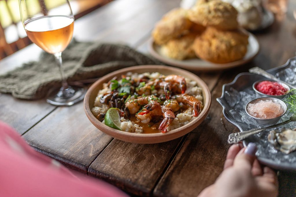 A glass of wine, a dish of shrimp and rice, an oyster platter and a plate of biscuits on a wooden table.