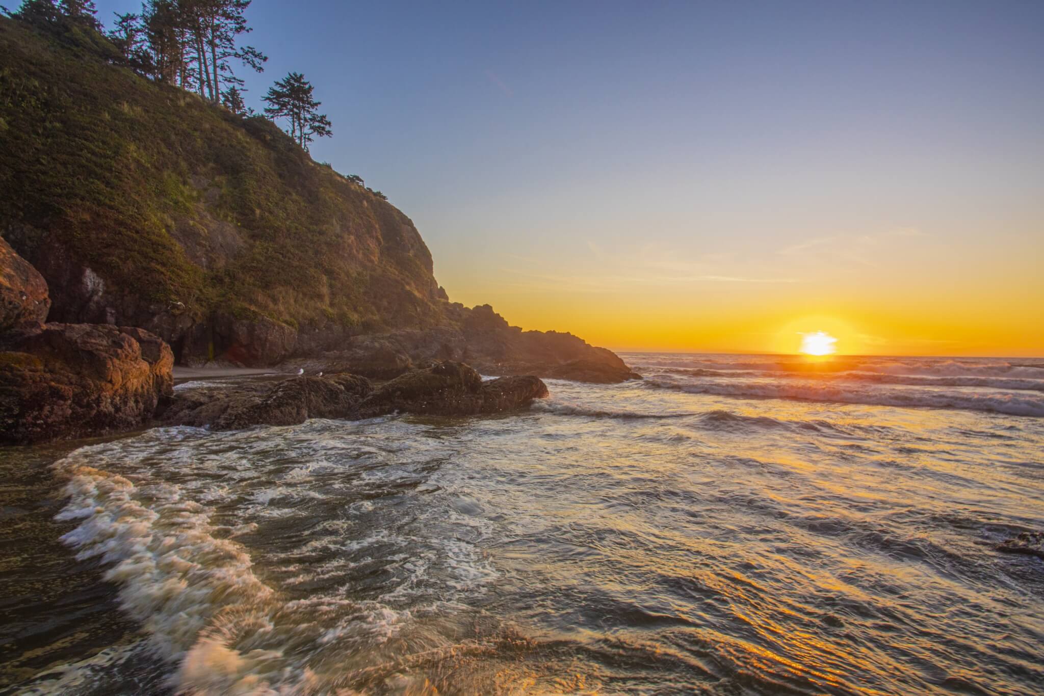 A grass-covered cliff beside the ocean and the sun setting over the water in the distance.