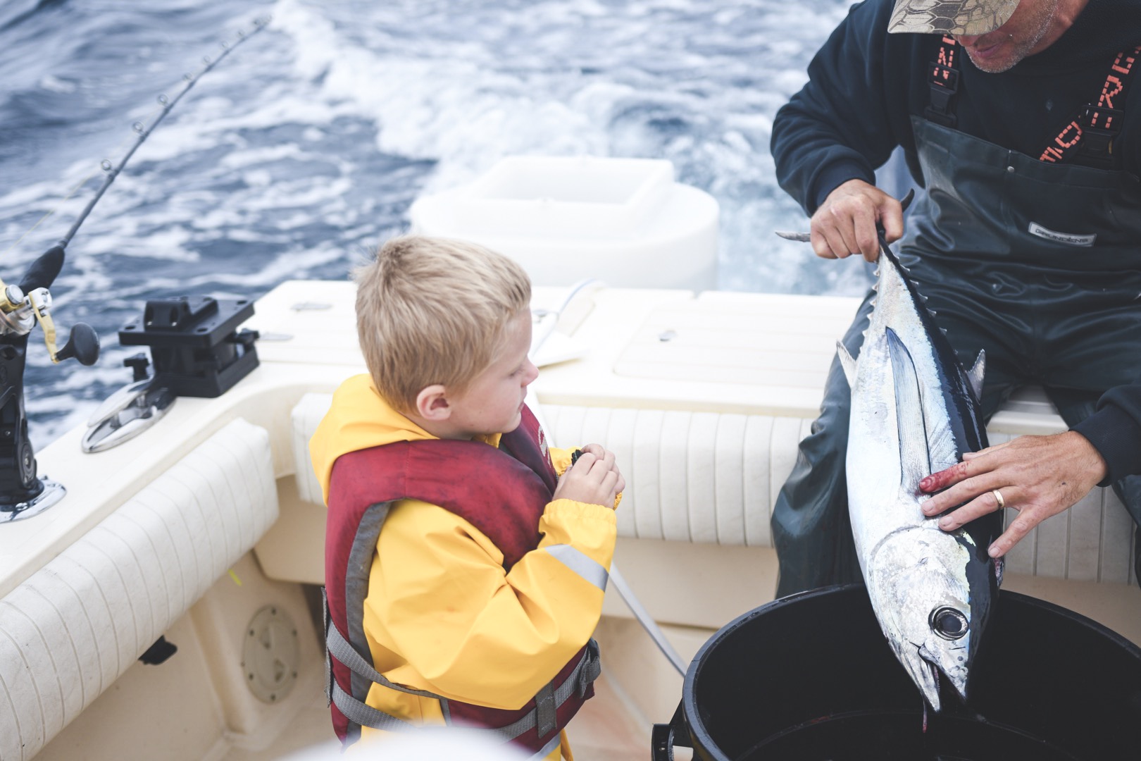 A child wearing a life jacket sitting in a fishing boat beside an adult holding up a large fish.