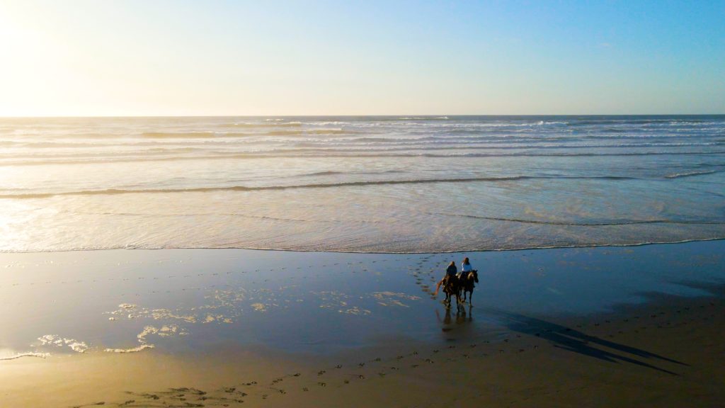 Two people riding horses on beach shore and rippling waves behind them.