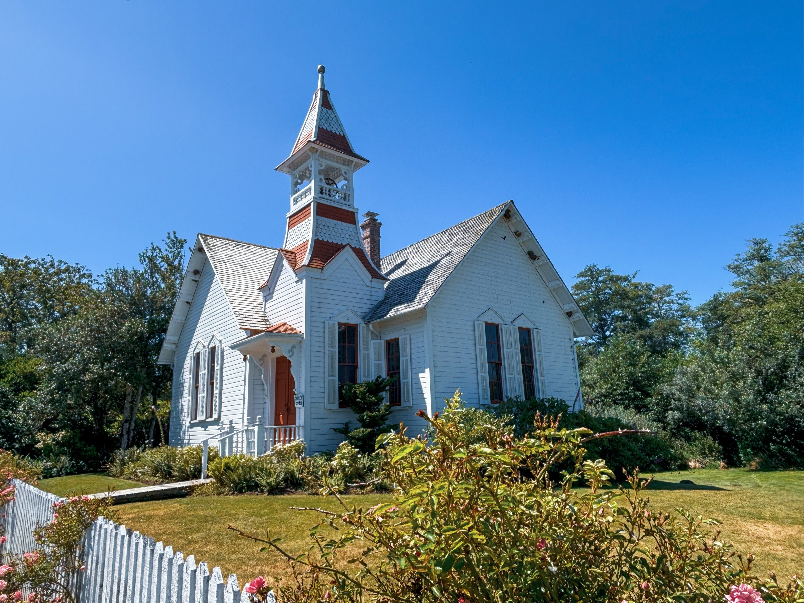 A white historic building with a small red- and white-striped tower and a forest of trees in the background.